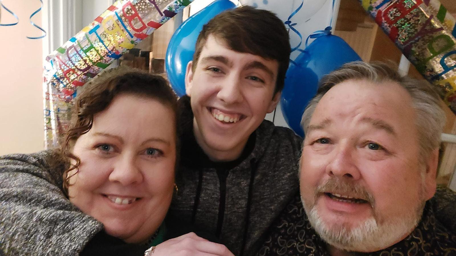 Michelle Mills a Christopher Mills yn gwenu gyda'u mab William rhyngddyn nhw. Mae baneri penblwydd hapus a balŵns yn y cefndir.