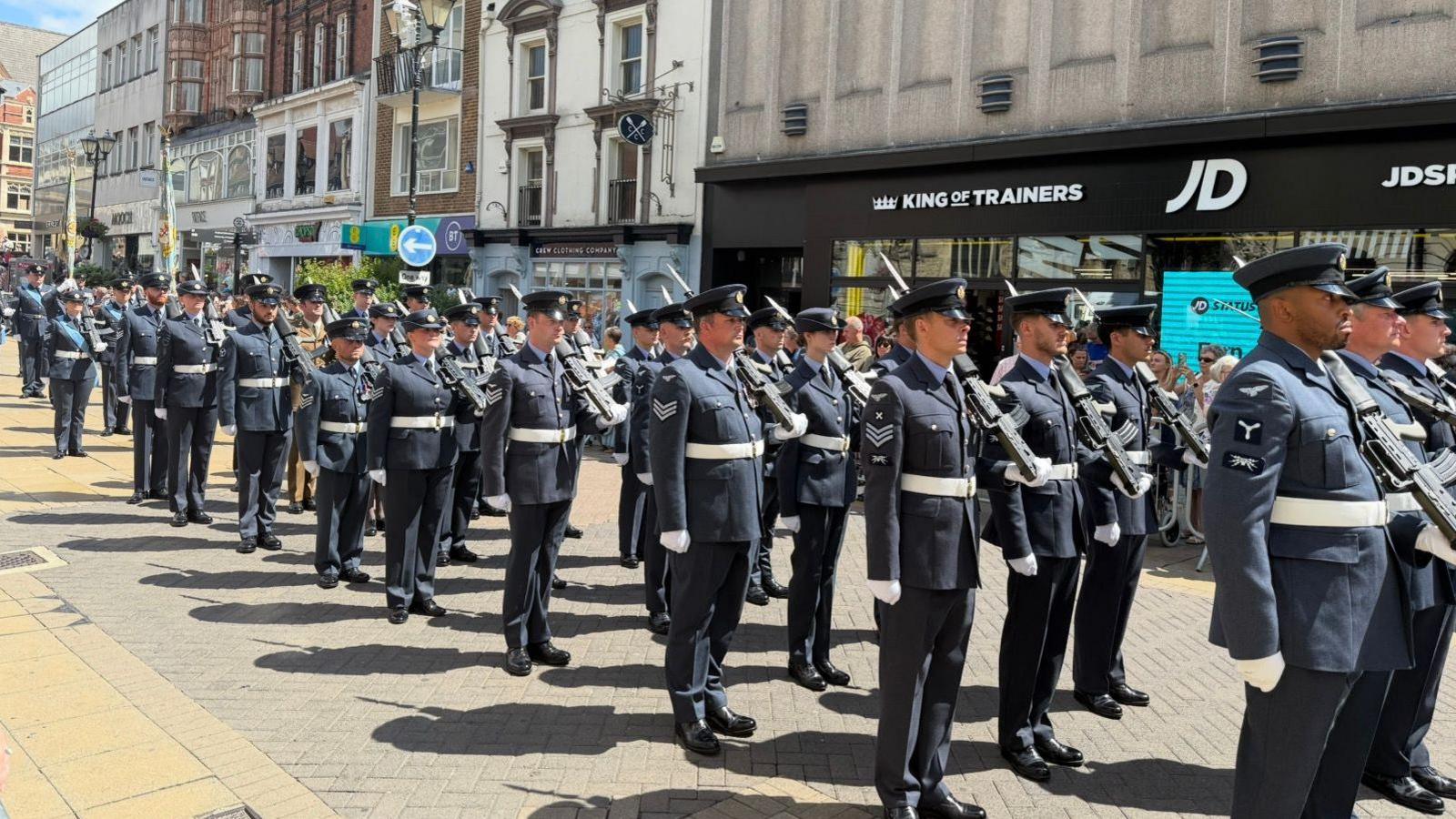 The photo shows officers wearing RAF uniform standing to attention. They're all wearing hats, and are carrying ceremonial swords. They're standing in the middle of a high street, which is filled with people spectating either side of them. 