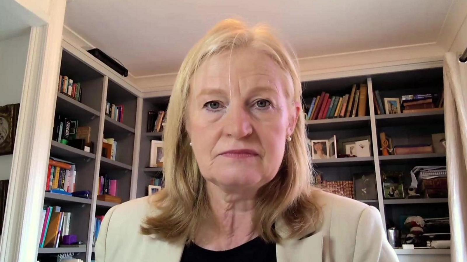 A woman in a beige jacket sits at a desk in front of shelves of books