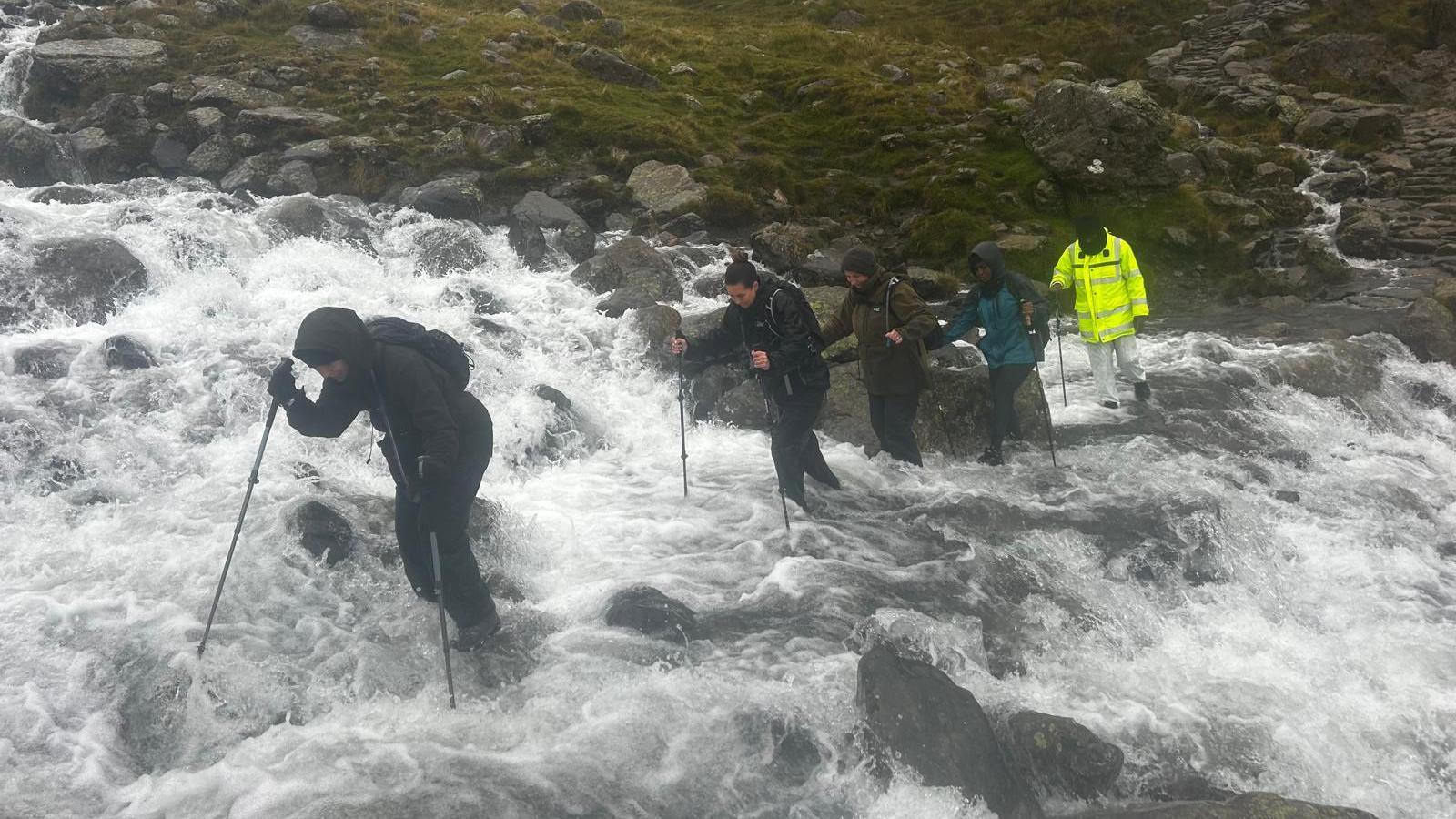 A group of five hikers attempting to cross a stony path which has flooded with water. The water has reaches about half-way up their shins. The group appear to be struggling through the water which has turned white and resembles rapids.