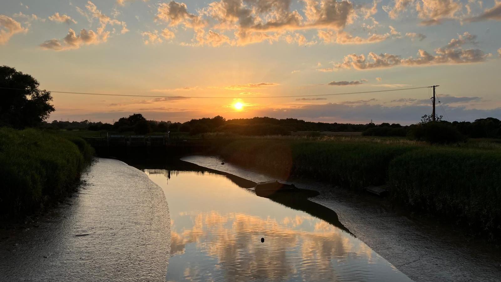 A waterway is pictured at RSPB Minsmere at sunset. The water in it is low, and the sun is close to the horizon. Trees and bushes are silhouetted against a pale sky