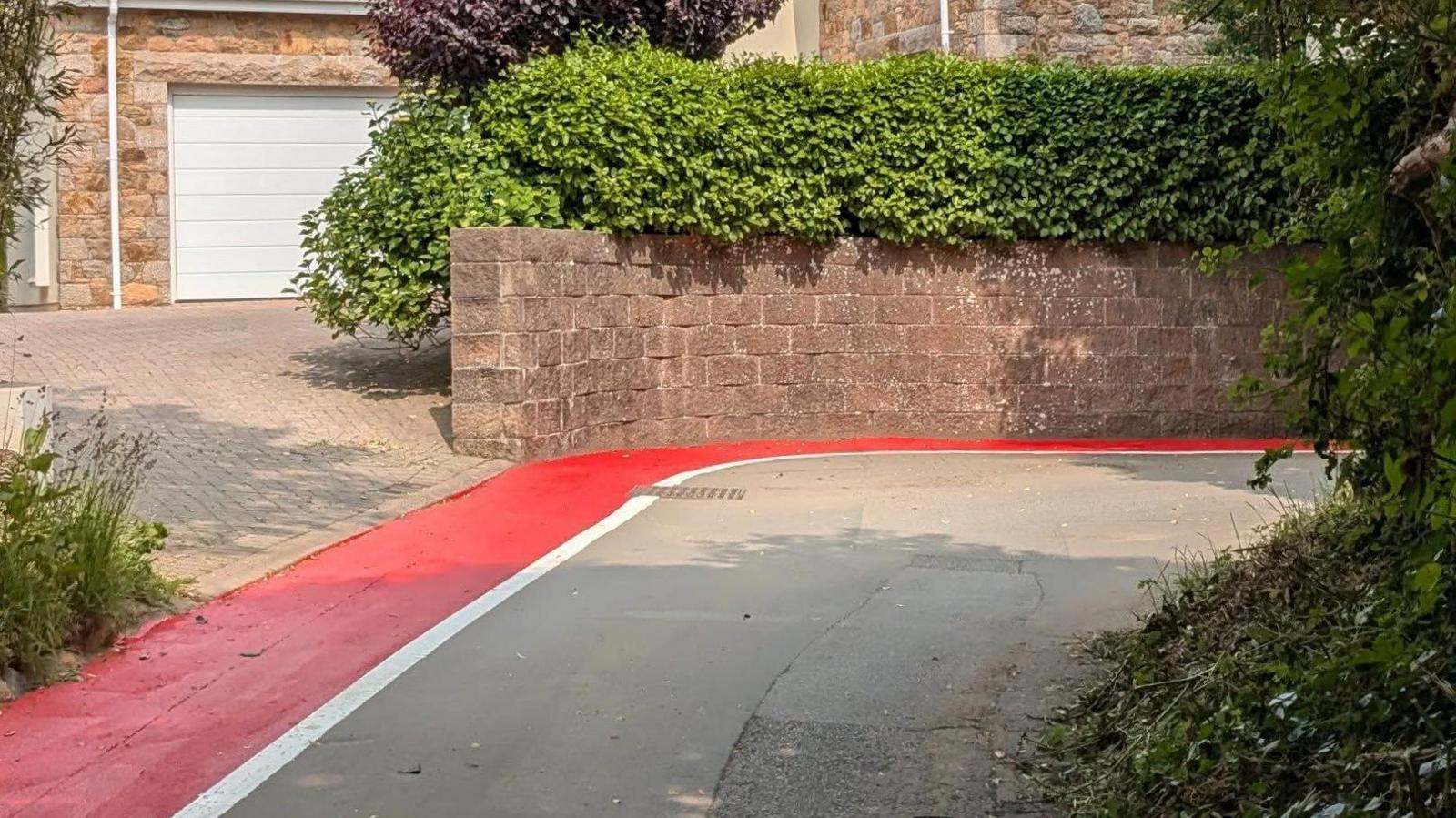 The picture shows a tight bend in a road with a bright red painted pedestrian walk way. The path has a white line to mark the pedestrian zone. There is a house and a driveway in the background with plenty of greenery.