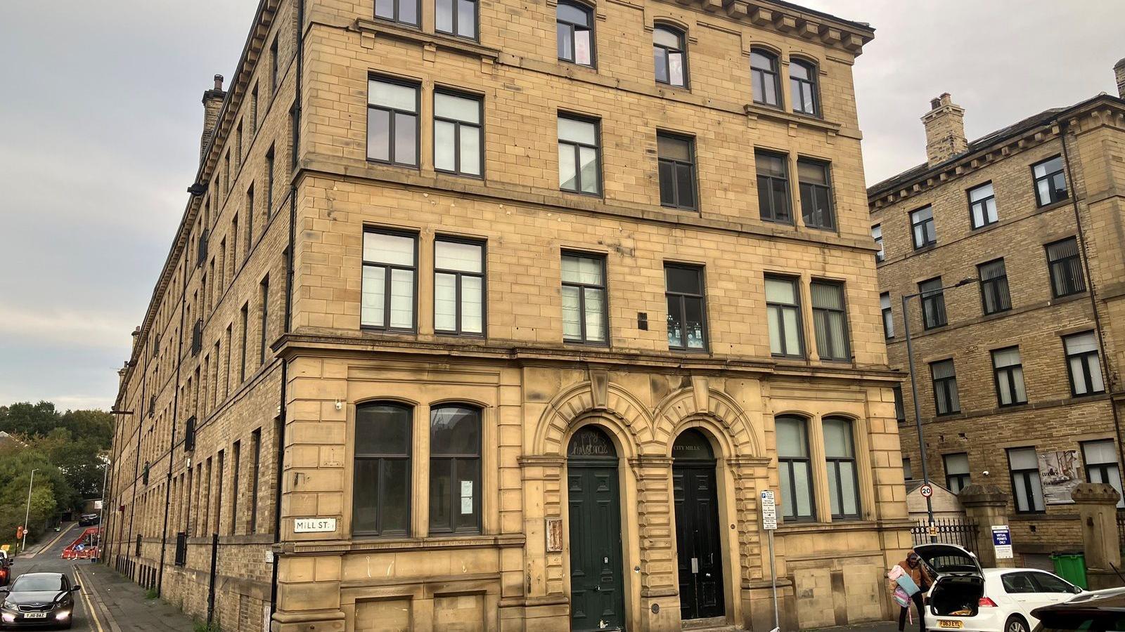 A large, four-story building constructed from light brown stone. Each floor has several rectangular windows framed in a dark material. On the ground floor, there are two prominent arched doorways fitted with dark green doors. A street sign on the corner of the building reads "MILL ST."
In front of the building, a cobblestone street runs along the edge, where several cars are parked. One white car has its boot open and a person is standing beside it. The sky is overcast. Adjacent to the main building, another similar stone structure is visible on the right.