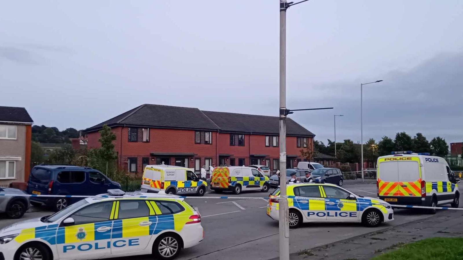 A number of police cars and vans are parked in front of a row of terraced houses, with a police cordon in place and several police officers and scene of crimes officers in attendance