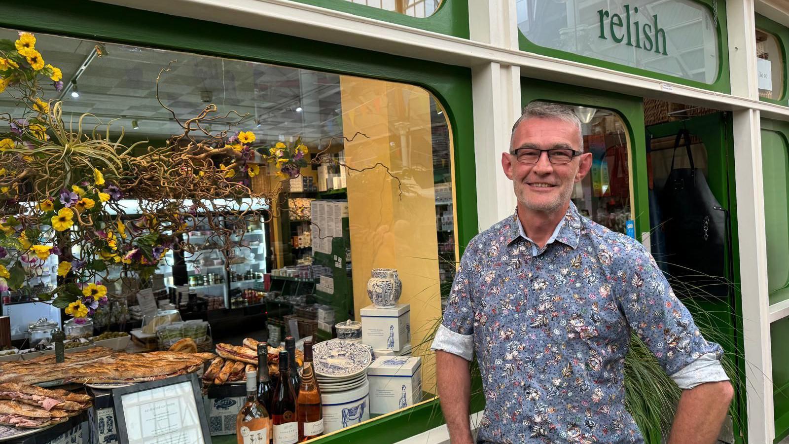 Romano - a man with glasses and grey/black hair wearing a rolled up shirt, blue base with floral accents, he is smiling and standing outside of Relish - the display window you can see blue and white china, rose wines, a range of ham sandwiches and a big bouquet of yellow and purple flowers