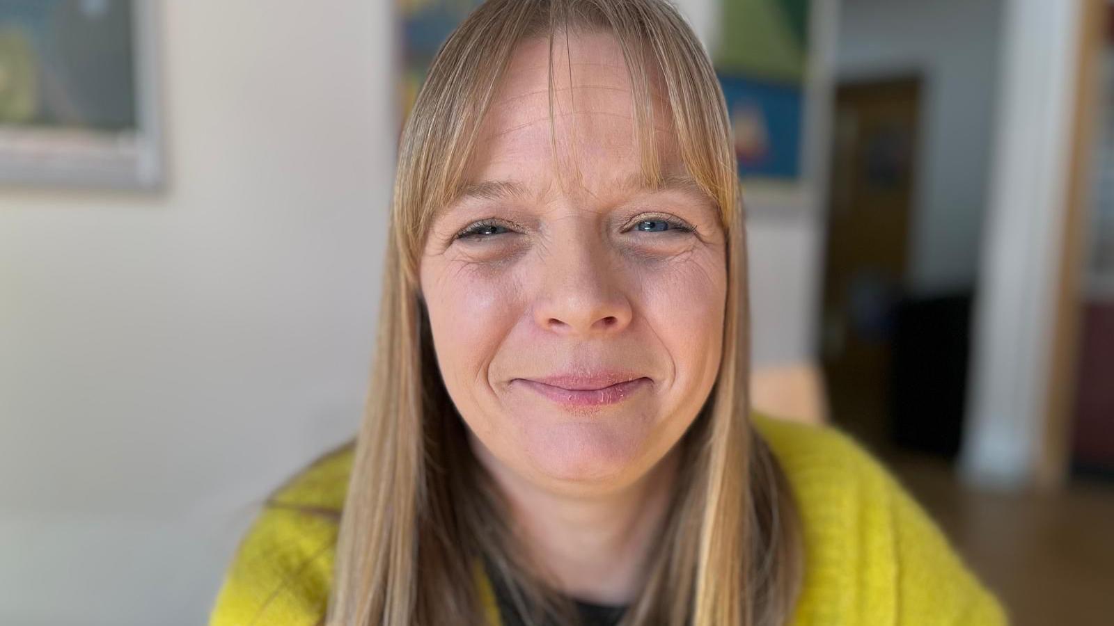 Emma-Jayne has long, blonde hair and is wearing a bright yellow, knitted cardigan, with a black top. The room behind her is out of focus.