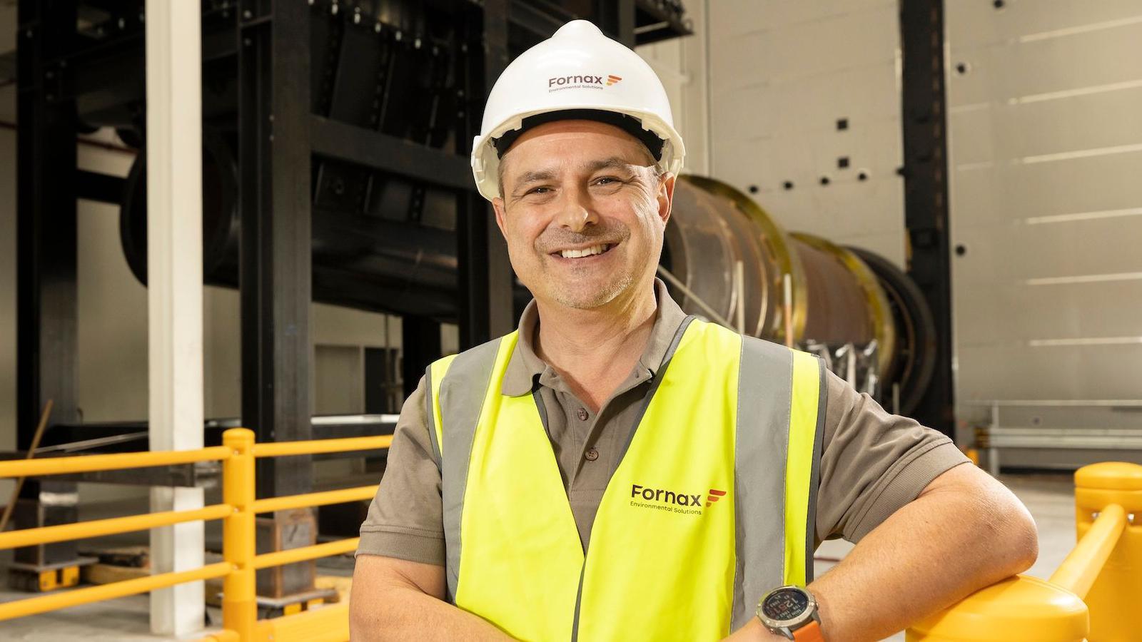 Ian Jones stands in the plant wearing a white helmet and high-vis jacket reading 'Fornax' on the right side. He has a watch on his wrist with an orange strap.