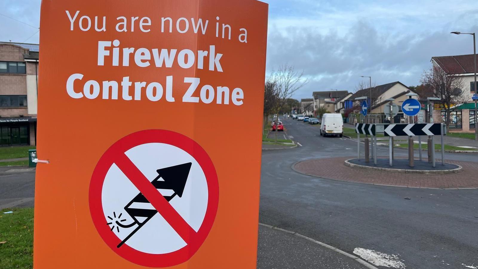 An orange sign warning people they are now in a firework control zone. The text on the sign is in white. There is a logo of a black and white firework on a white background with a red circle around it and a red strike through the middle.