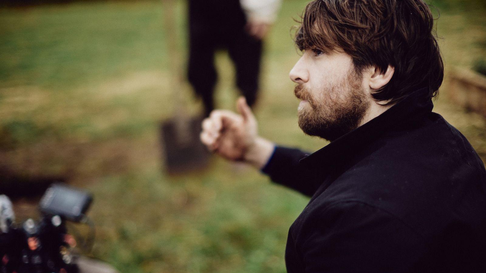 A brown haired man with a beard, wearing a dark coat in a field. He looks towards a camera operator who is slightly out of shot, giving them a thumbs up. An actor holds a shovel in the background.