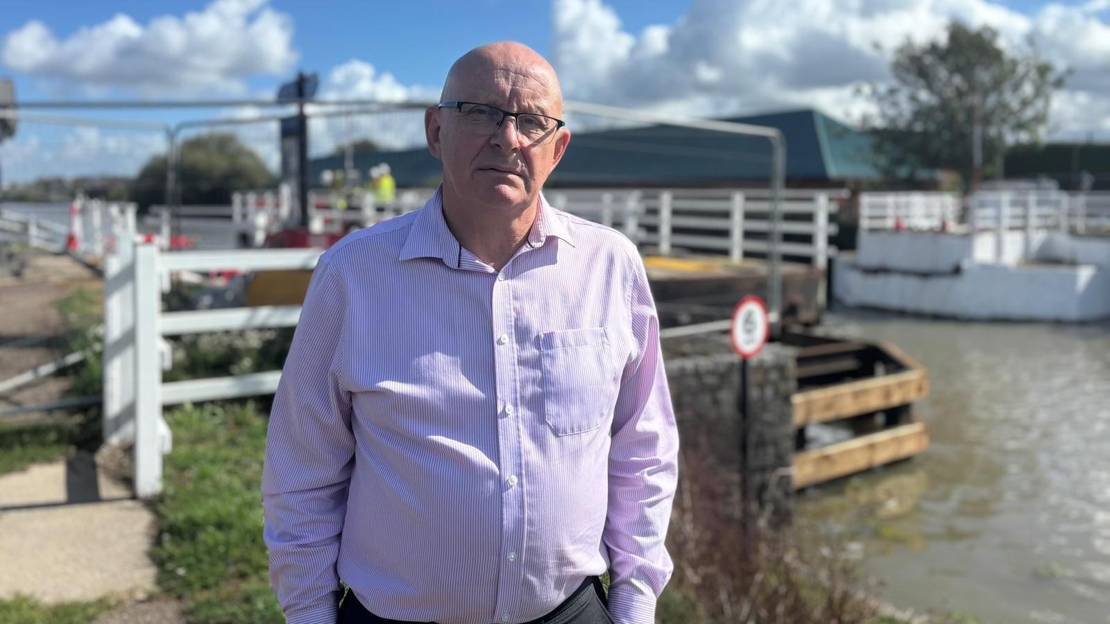 A man in a pink-purple shirt and grey trousers stands in front of Netheridge Swing Bridge. He is wearing glasses and looking directly at the camera.