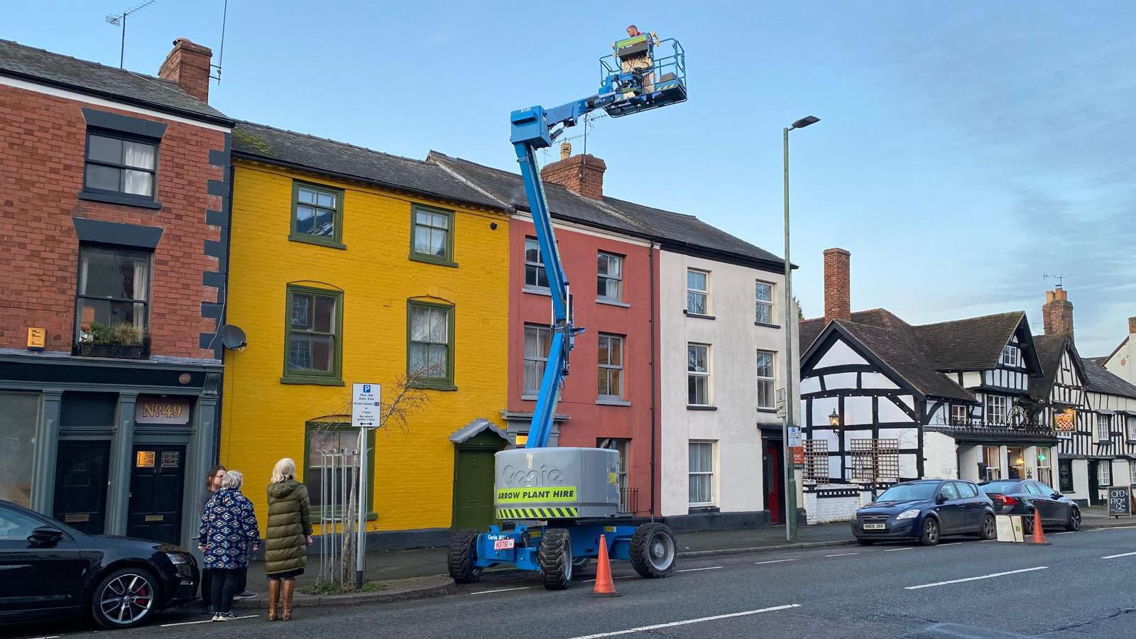 A row of terraced houses with brick, yellow, terracotta and cream frontages. On the street in front of them is a blue cherry picker with a man in the basket at roof height. 