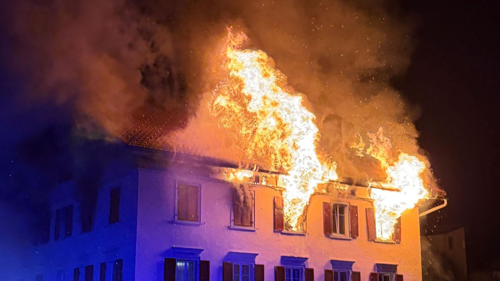 Large flames come out of the windows in the top floor of a building at night.