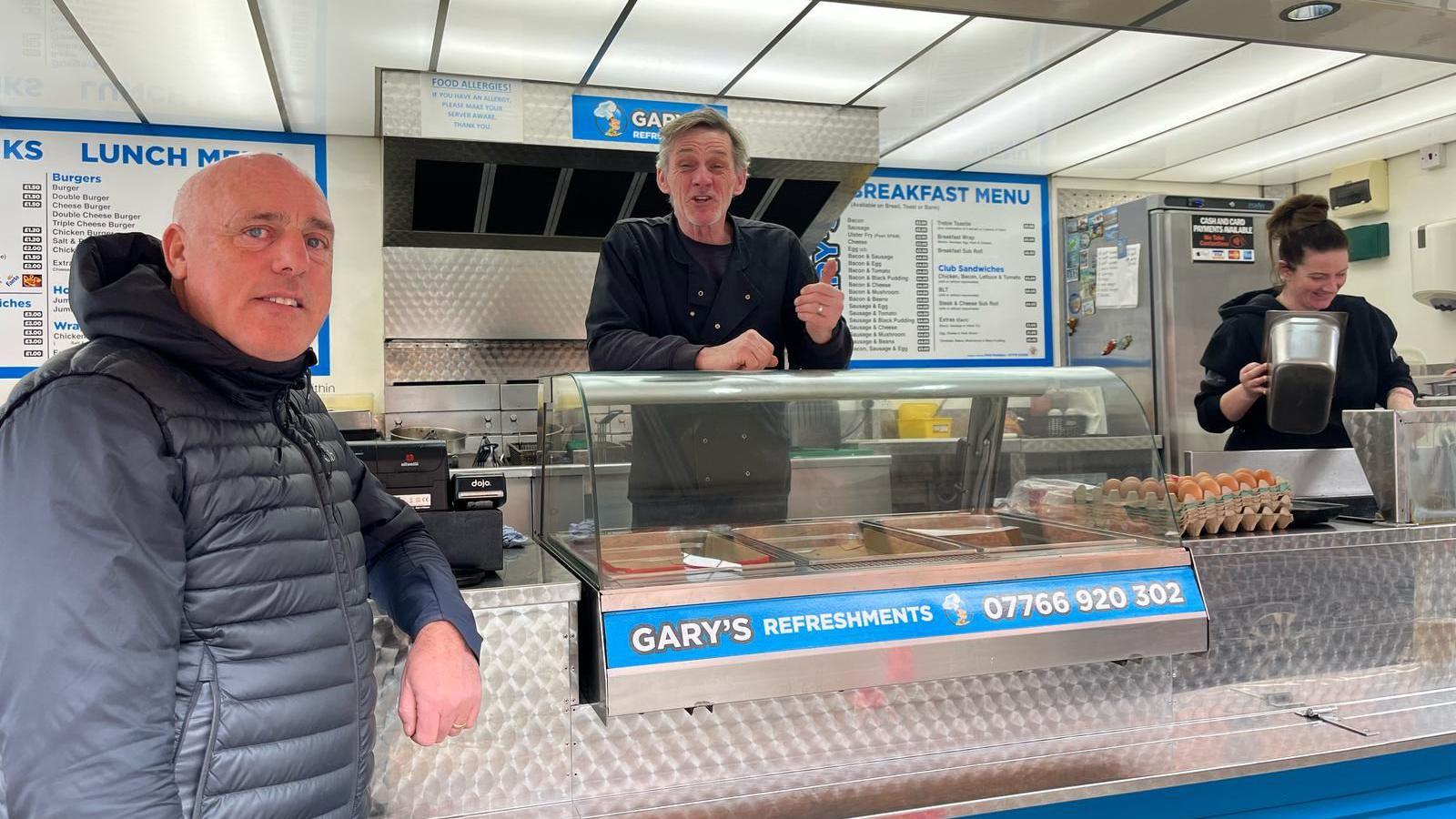 Carl Ennis, who has a bald head and wears a black bubble coat leans on the counter of a food fan behind which stands Gary Buckley, who has grey hair, and on the right a woman is preparing food.