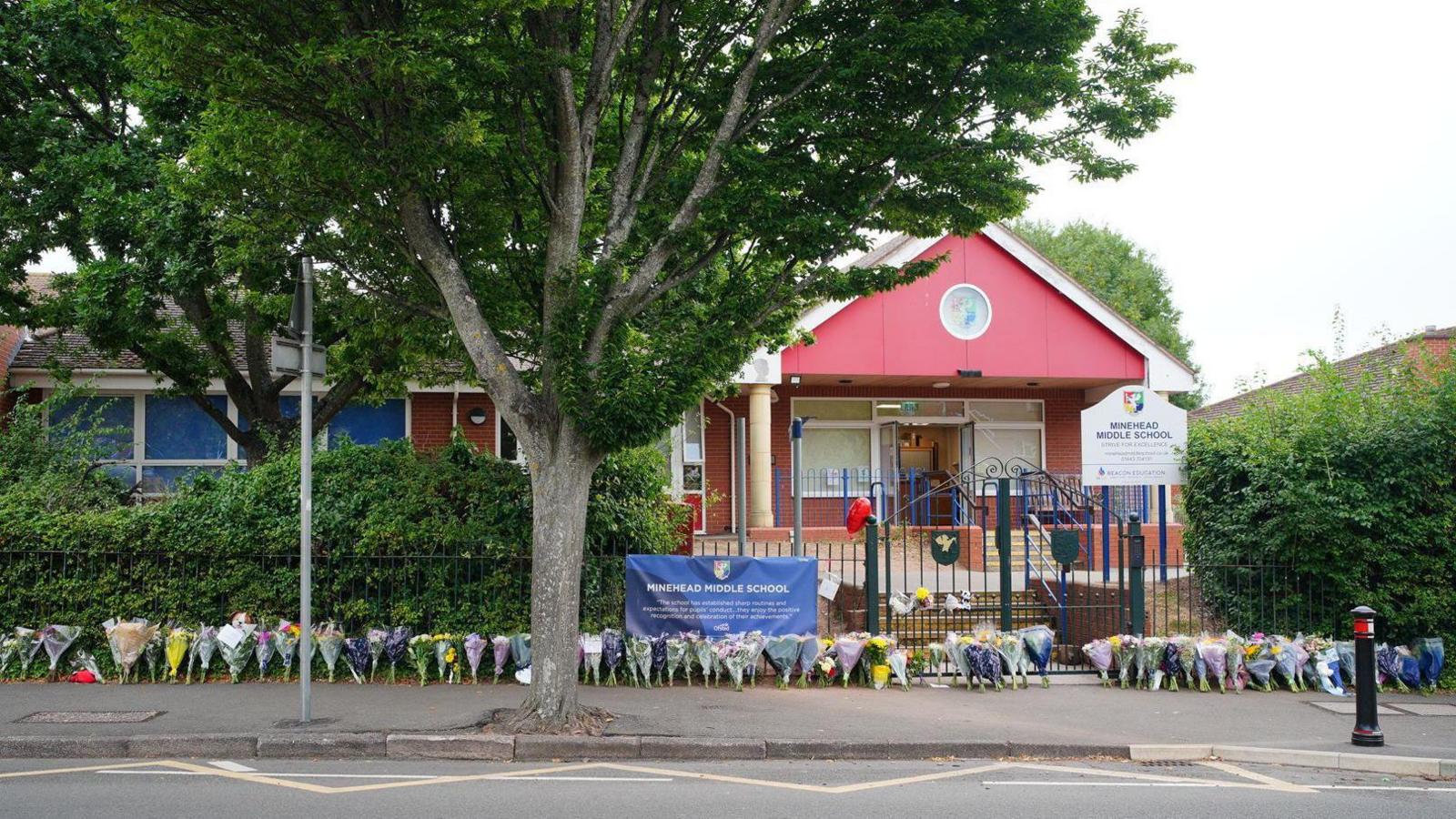 Minehead Middle School. The building is red and there is a green metal gate at the entrance. The street in front of the school is lined with bouquets of flowers left in memory of a pupil who died.