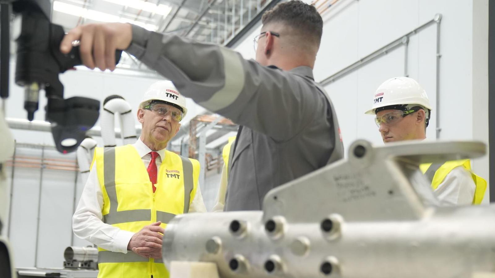 A man in a hard hat and safety jacket looks thoughtfully as another man in a grey boiler suit points at a machine