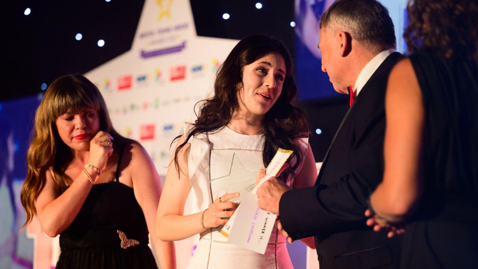 Aliese has long dark hair and is wearing a white dress as she stands on stage with her prize. She is talking to a man on stage.