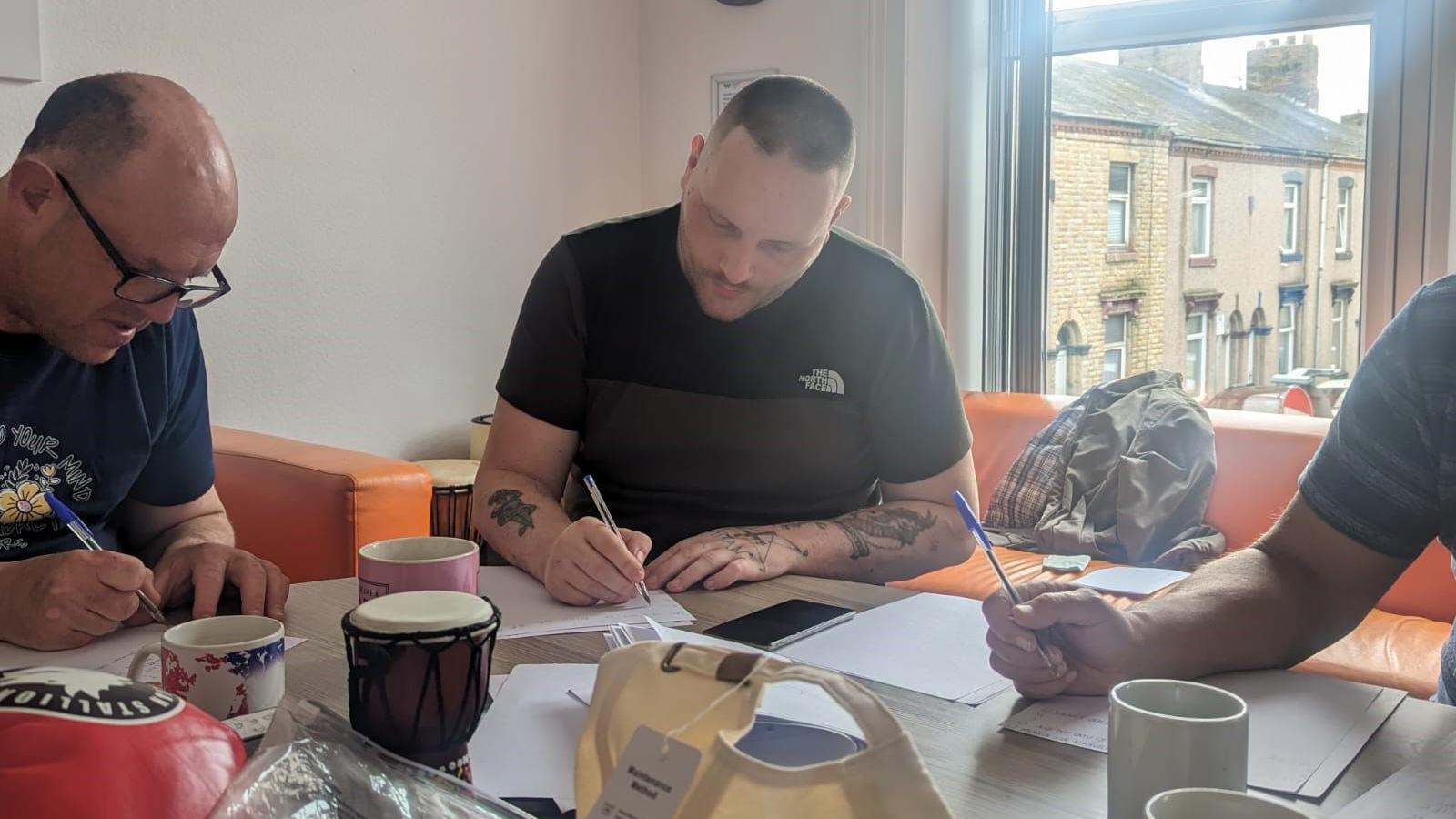 Men sitting round a table writing, with Barrow terrace housing visible out of the window