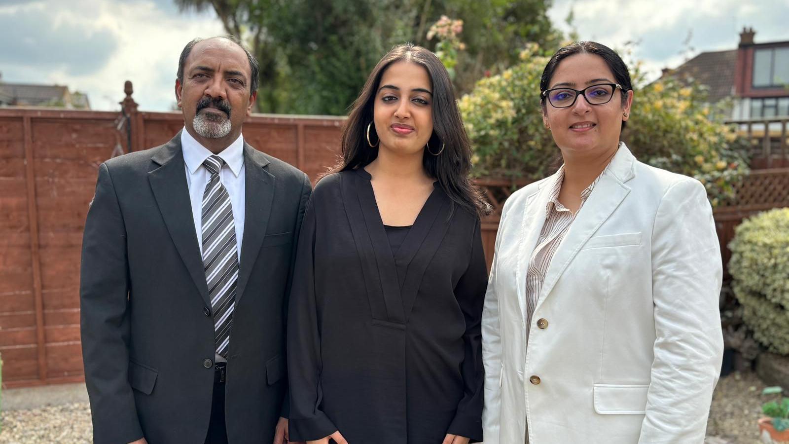 Ms Cheema, a woman with long dark hair and wearing a black top, standing between her parents. Her mother on the right is wearing glasses and a white blazer and has her dark hair tied in a low bun and her father on the left has a greying beard and is wearing a black suit with a stripy tie. They're standing in a garden with a wooden fence behind them. It's a cloudy day.
