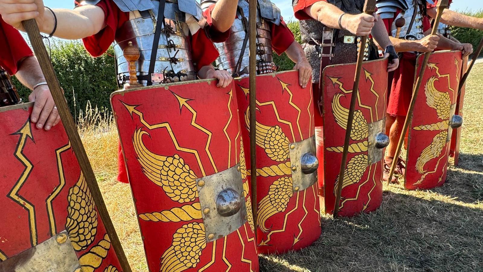 A line of bright red Roman recreation shields, with gold centres and yellow markings on them. They are held by people dressed as Roman soldiers. 