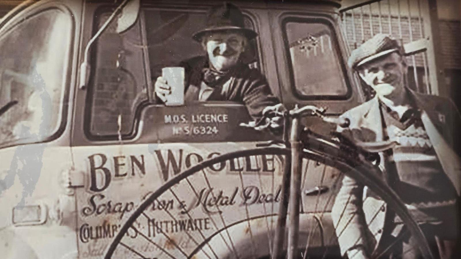 A black and white photo shows a man in a hat holding a cup out of the window of a truck. Next to it is a man in a flat cap and a penny farthing bicycle.  