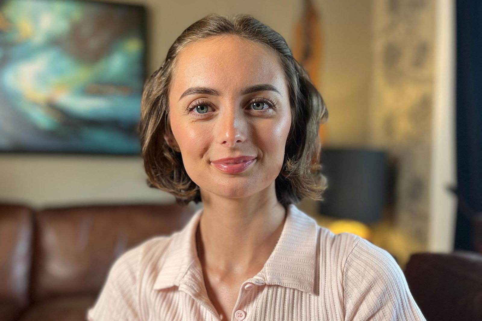 Person seated indoors on a brown leather sofa, wearing a light pink ribbed collared top with a clip-on microphone attached. A framed abstract painting hangs on the wall in the blurred background, with soft indoor lighting and shallow depth of field creating a professional interview-style setting.