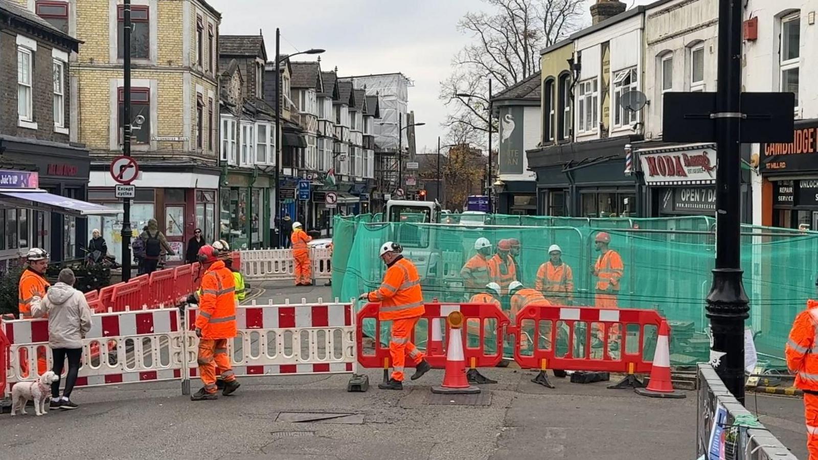 Red and white barriers cross a road with a green transparent fence behind it. A number of men wearing orange high-visibility work suits are present.