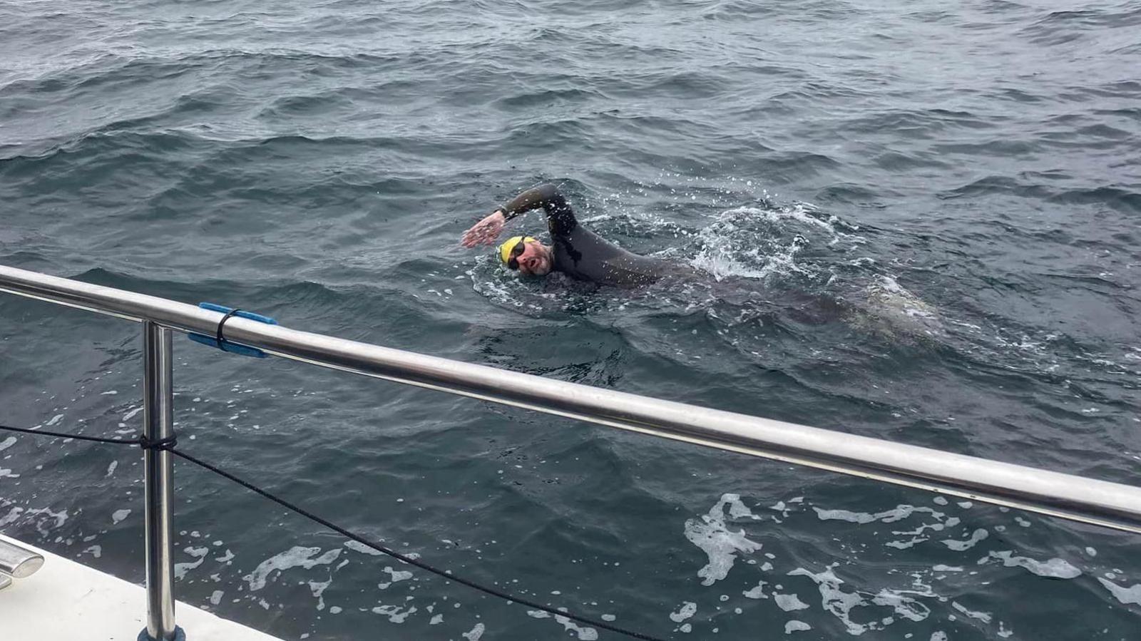 One of the swimmers in the sea. He's wearing a yellow swim cap and black goggles and is in a wetsuit. You can see the silver metal railing of the boat.