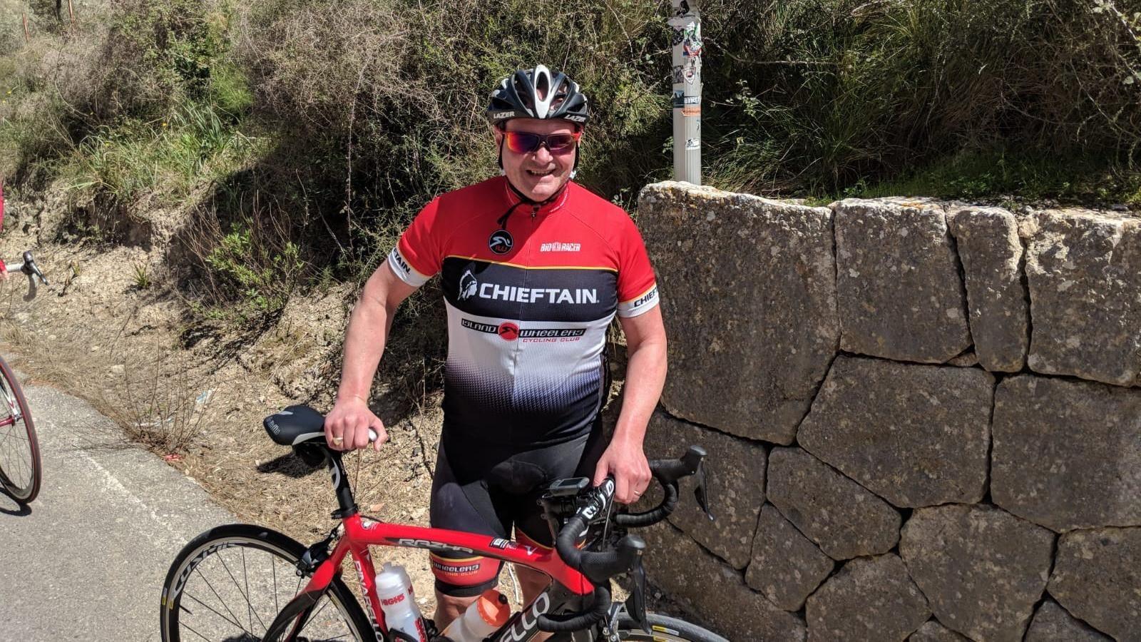 Paul McAdam wearing a cycling speed suit which is red black and white with 'Chieftain' written across the front of it. He is holding a red bicycle upright standig in front of a stone wall.  