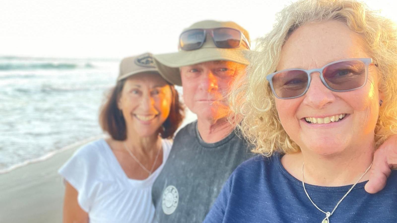 Jess wearing glasses and a blue top, with curly blonde hair, standing on a beach, with Alistair and his wife Suzie standing behind her.