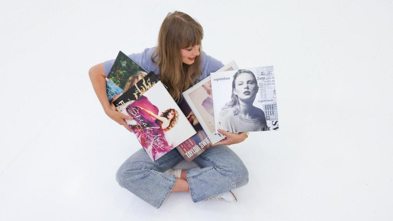 Taylor Swift sits cross-legged on the floor, holding copies of her first six albums
