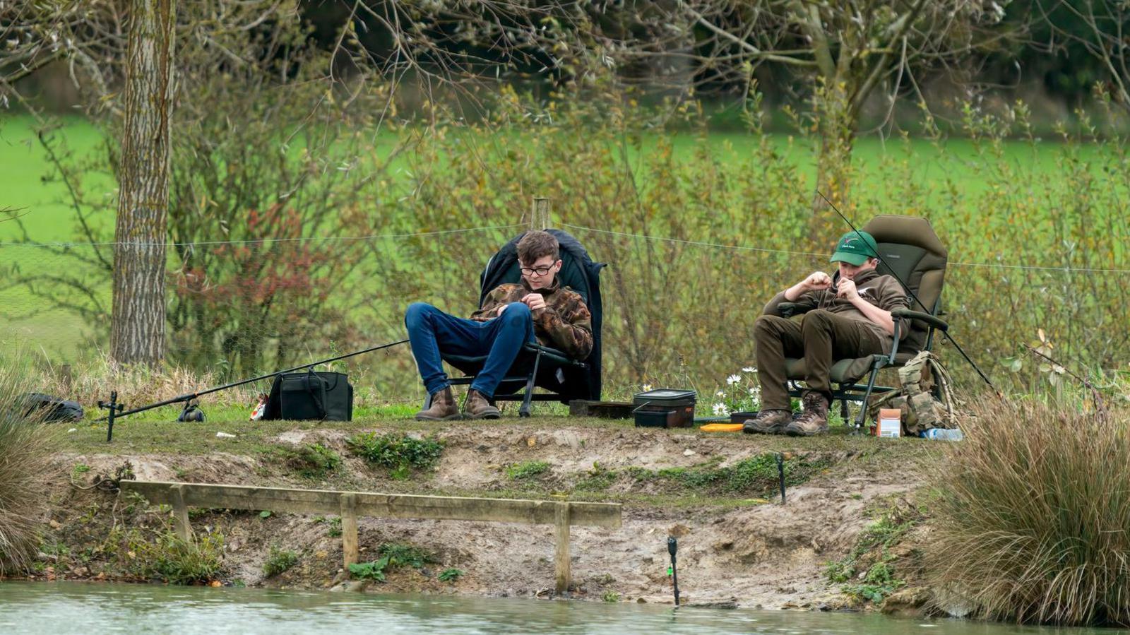 Two teenage boys sitting by a lake in camping or fishing seats, their fishing rods in the water. They are in dark clothing. The backdrop is a green field and foliage.