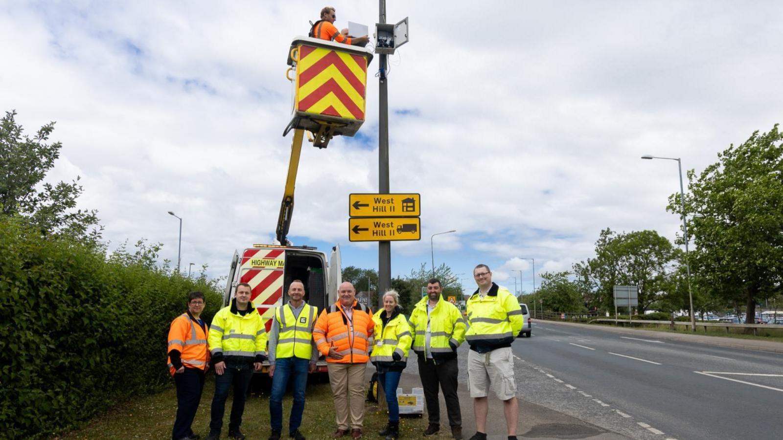 Seven people standing in a line wearing yellow and orange high-vis jackets and looking at the camera. They are standing on a grass verge next to a road. A highways van is behind them with a crane, and a man standing in the tub at the top.