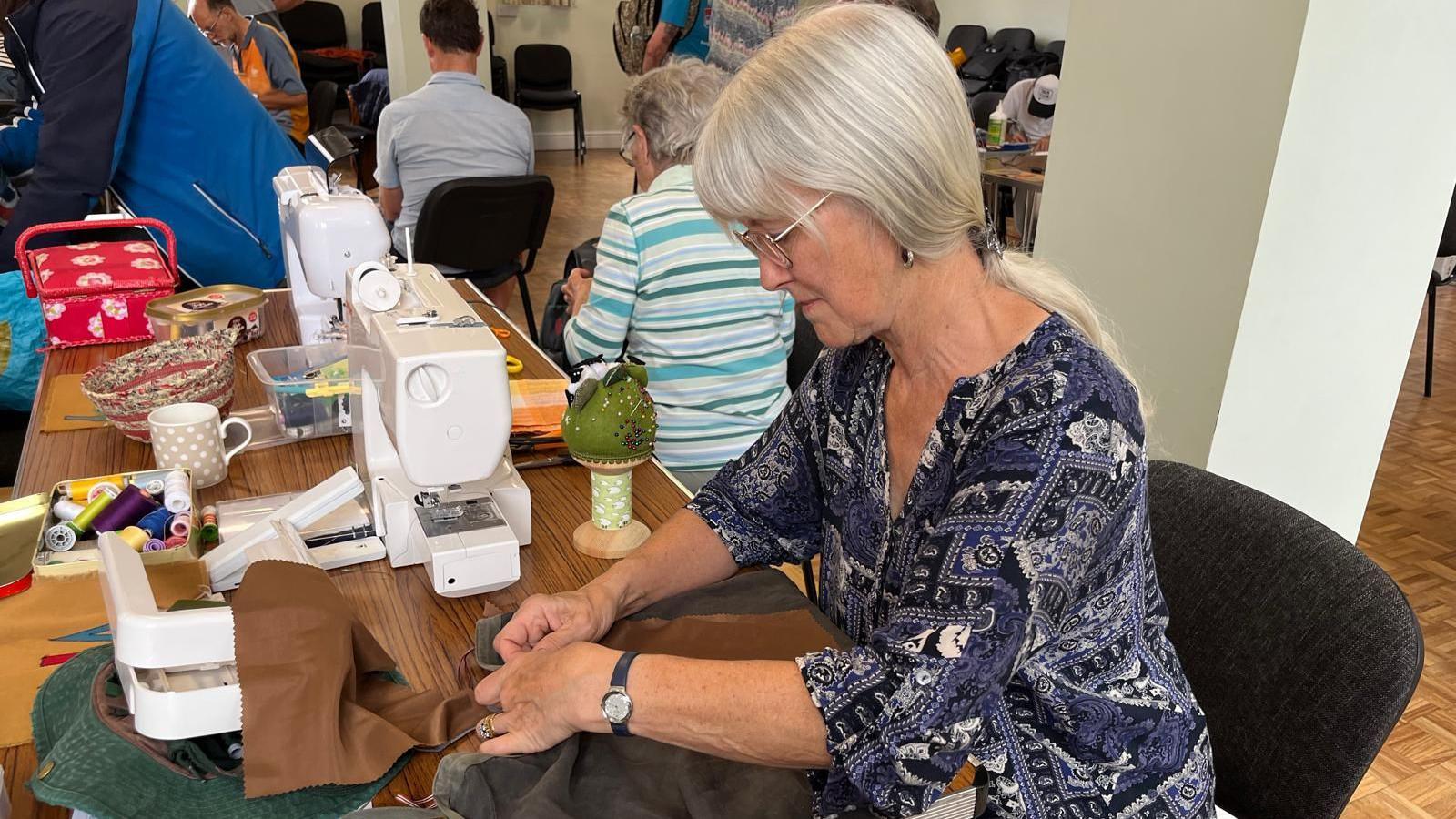 A woman with long grey hair in a ponytail holds a grey item of clothing. She sits in a black chair, while working on the item which is on a long brown table. Two sewing machines are on the table.