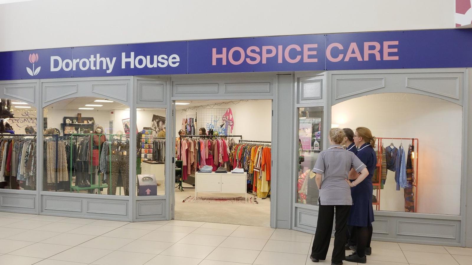The frontage of a Dorothy House shop inside a shopping centre. Three women in nurses uniform stands chatting outside. There are lots of colourful garments on rails to be seen through the large glass panes.