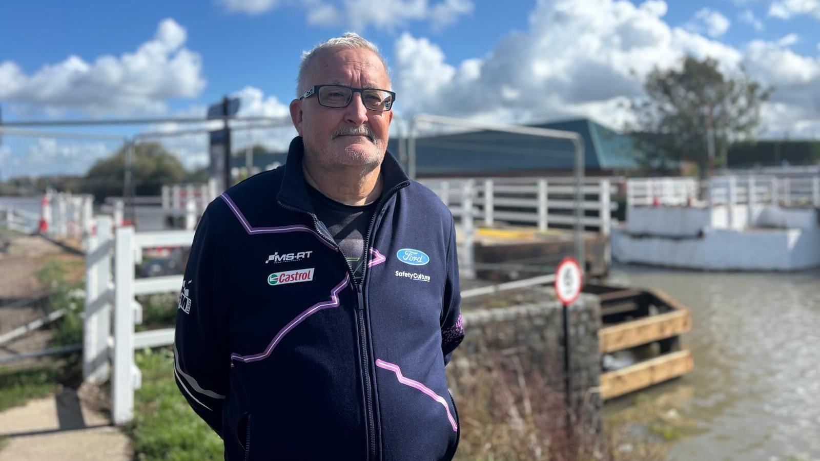 A man with grey hair, glasses, and a moustache wearing a navy jacket with Ford and Castrol logos looks at the camera with the canal and bridge behind him.