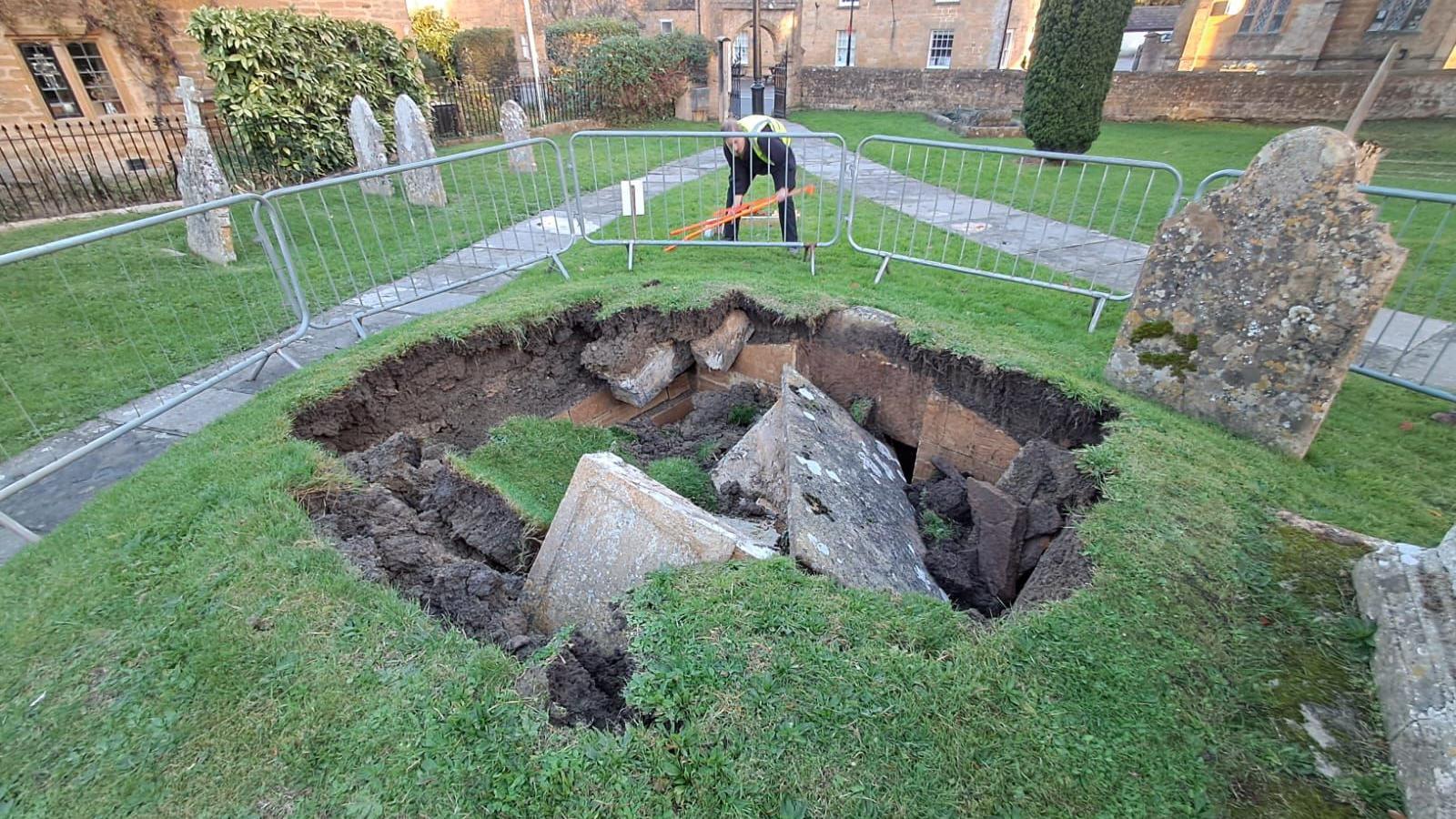 A stone box tomb which has collapsed into a large hole in the ground, through the soil and into the crypt below, a small stone home buried underground. There are metal barriers around the hole and a man in a high-vis jacket. There are other gravestones very close to the edge of the hole, which is about 12ft (3.6m) wide.