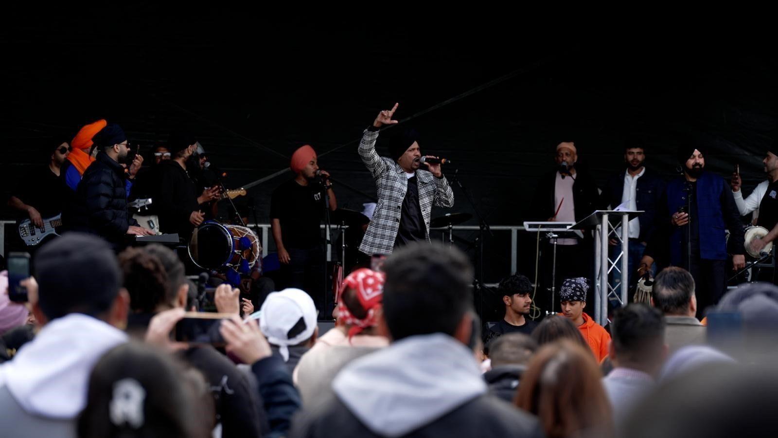 A man in a turban stands on a stage with a microphone in one hand and his other hand in the air. Around 10 other men stand on stage with various instruments or microphones. They stand in front of a crowd, who are watching the stage.