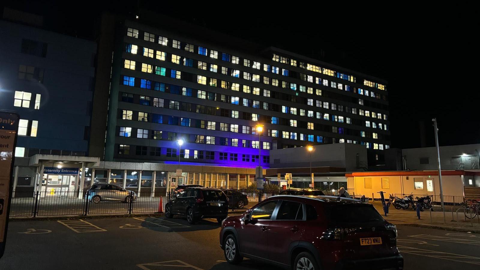 A car park at night at a hospital. A few cars can be seen in the car park. An "Entrance" sign is at the front of the car park with a large building behind which has dozens of square windows