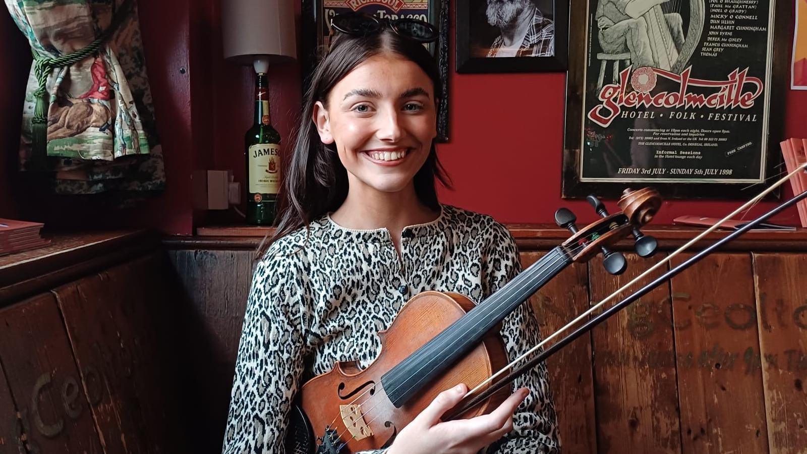 Rosie is sitting in a bar holding her fiddle She's smiling at the camera.