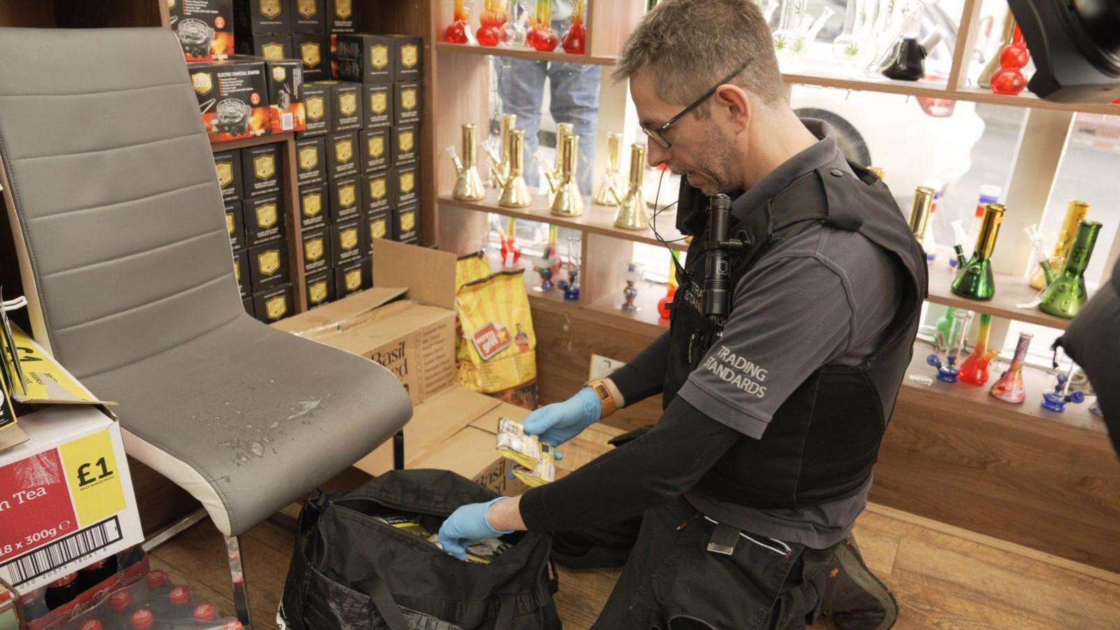 A Trading Standards officers wearing black and grey clothing and blue latex gloves rummages through a black bag in a shop to search for illegal tobacco products