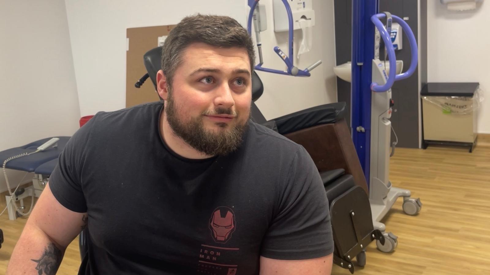 A man is sat down in a hospital setting. Behind him there is a mobility chair, a mobility crane, a hospital bin and a therapy bench in a white room. The man has dark hair and a full dark beard. He is wearing a black t-shirt with a red logo.