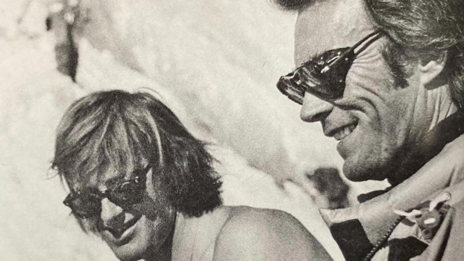 Martin Boysen is sitting on the side of a snowy next to the actor Clint Eastwood. Both men are wearing sunglasses and are smiling in this candid black and white image. 