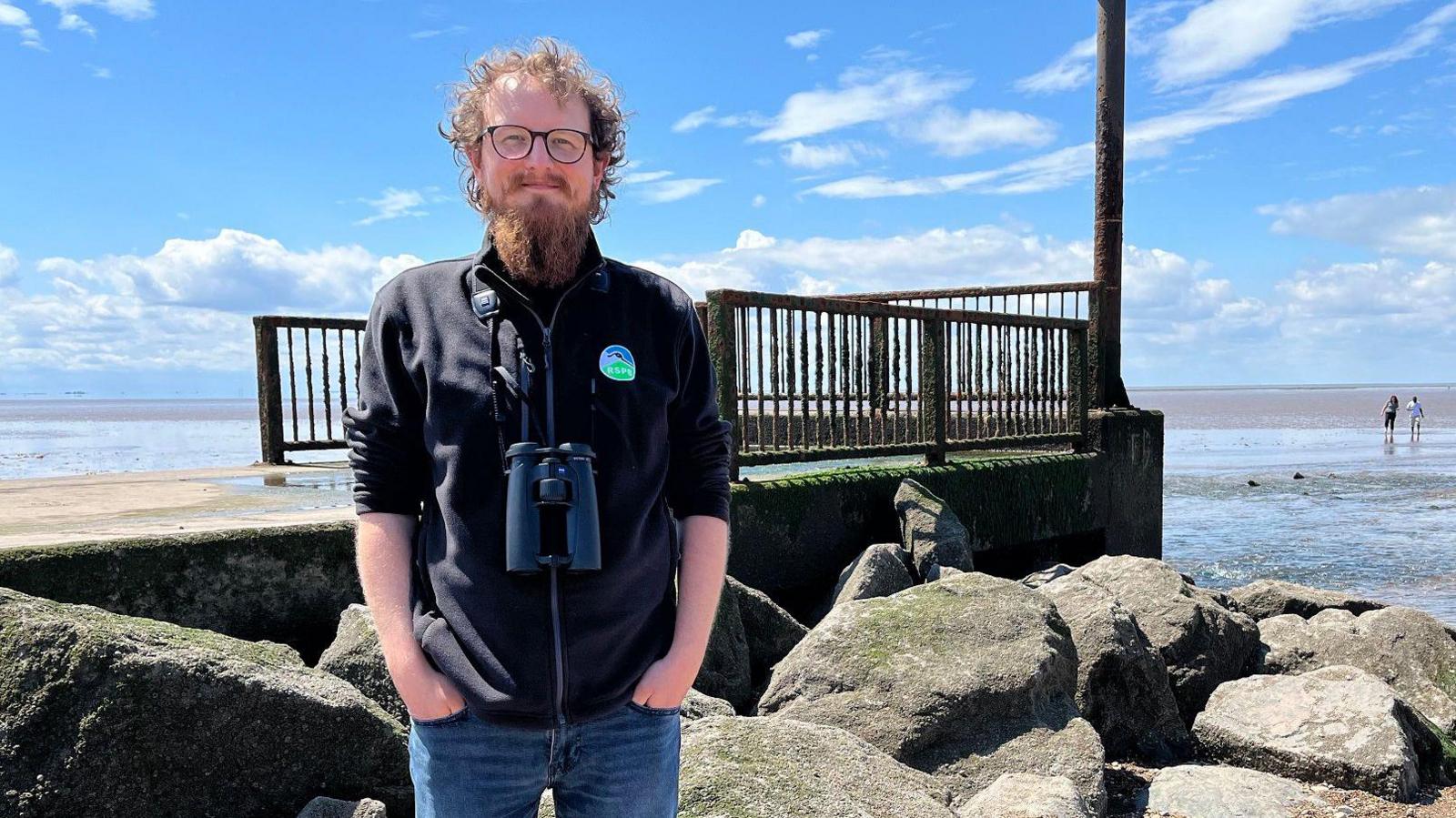 Ed Tooth from the RSPB stands in front of rocks with the sea in the background. He has red curly hair and wears glasses and has a pair of binoculars around his neck. He wears a black gilet and blue jeans.
