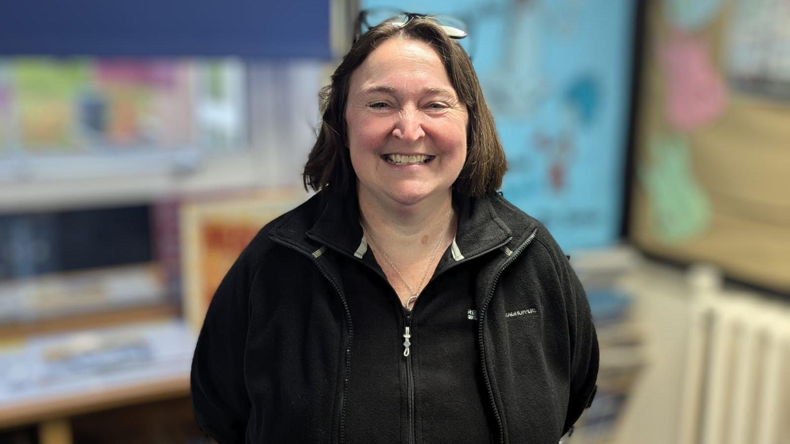 A woman with long brown hair, wearing a black zip-up jacket, smiles at the camera. 