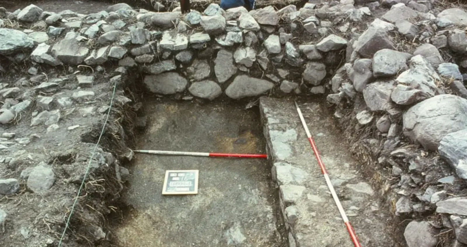 An exposed area of stone-built walls and a floor. There are red and white measuring poles laid out on the ground. 