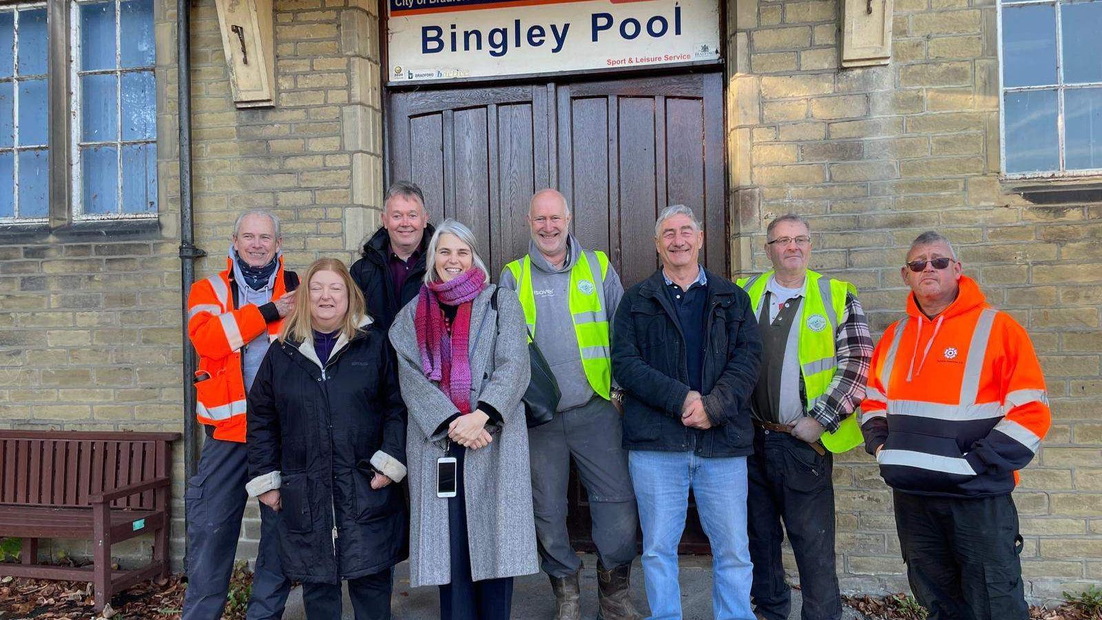 A line up of smiling men and women outside the wooden door entrance to Bingley Pool