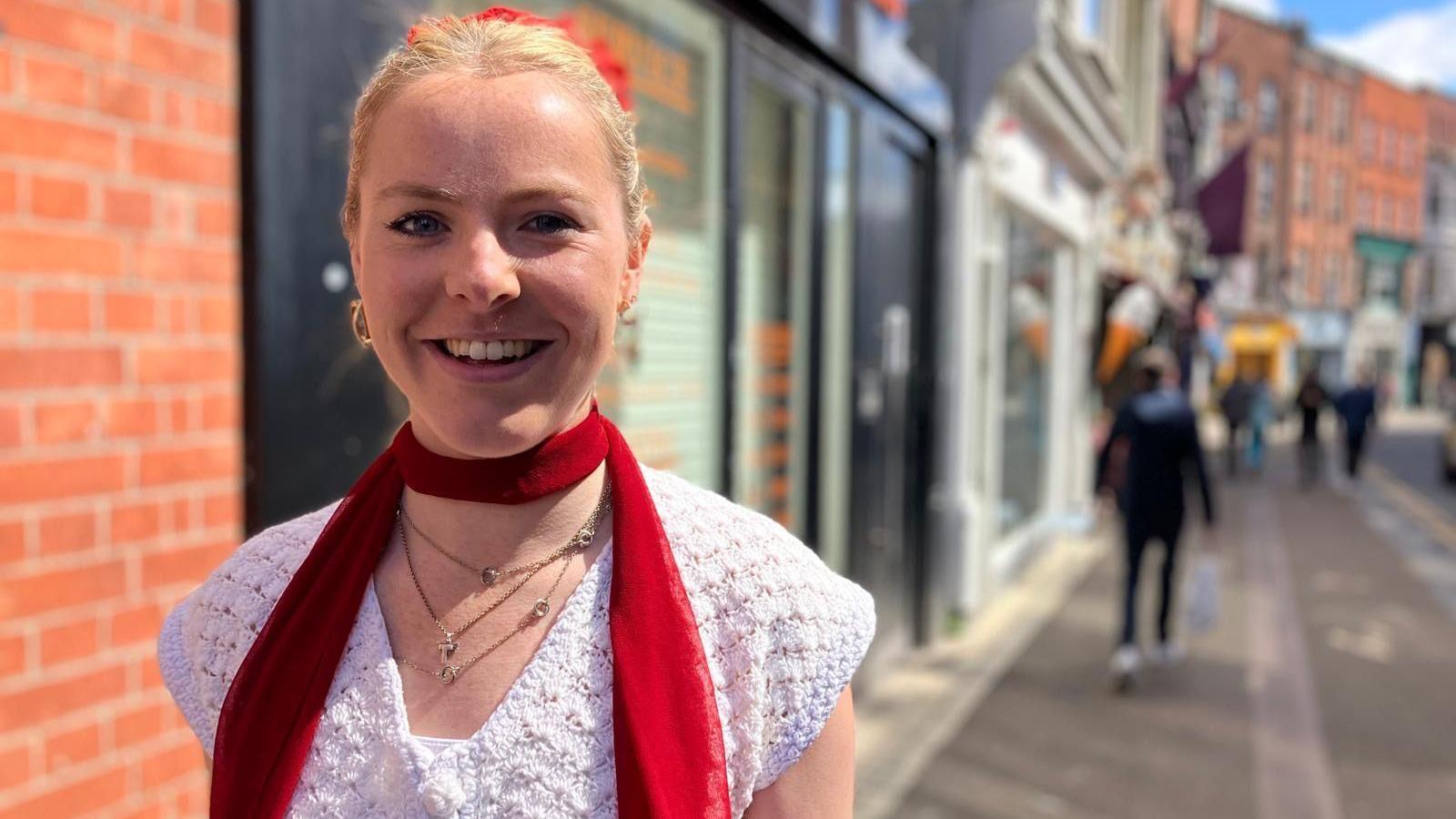 A woman has blonde hair, scraped back from her face in a red bobble. She is smiling into the camera. She wears a white top and a skinny red scarf.