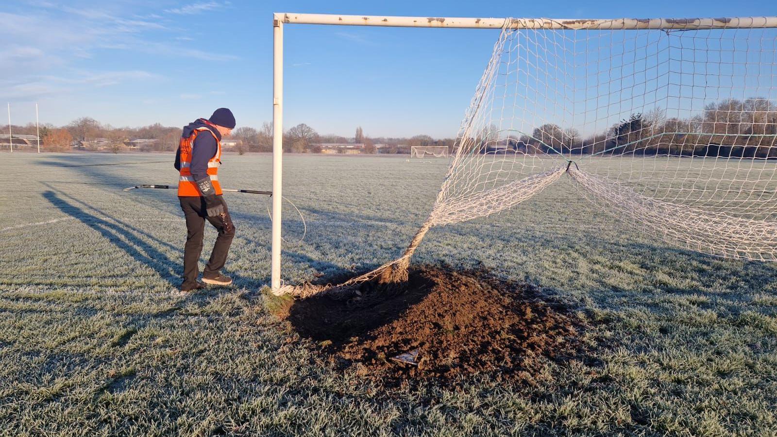 A football goal with a large brown hole dug in the ground under it. A man approaches with a high-vis jacket. The grass is covered in frost.