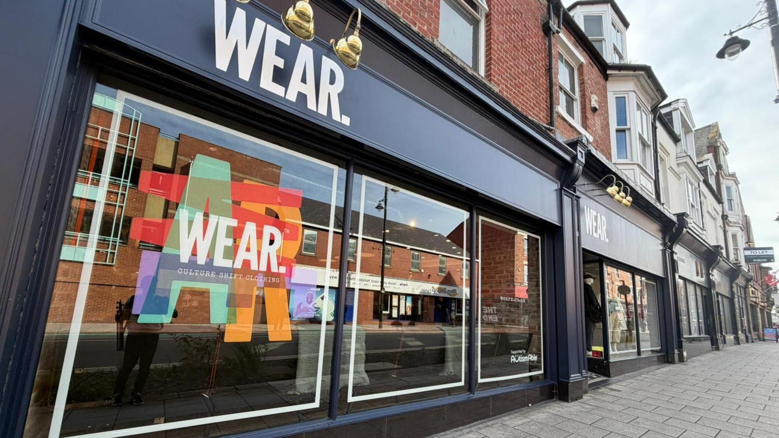 The outside of the Wear shop, which is painted in a very dark blue and has a number of gold-coloured lamps lighting up the words Wear below. There is one large multi-coloured logo, which says Wear, being displayed in one of the large front windows.