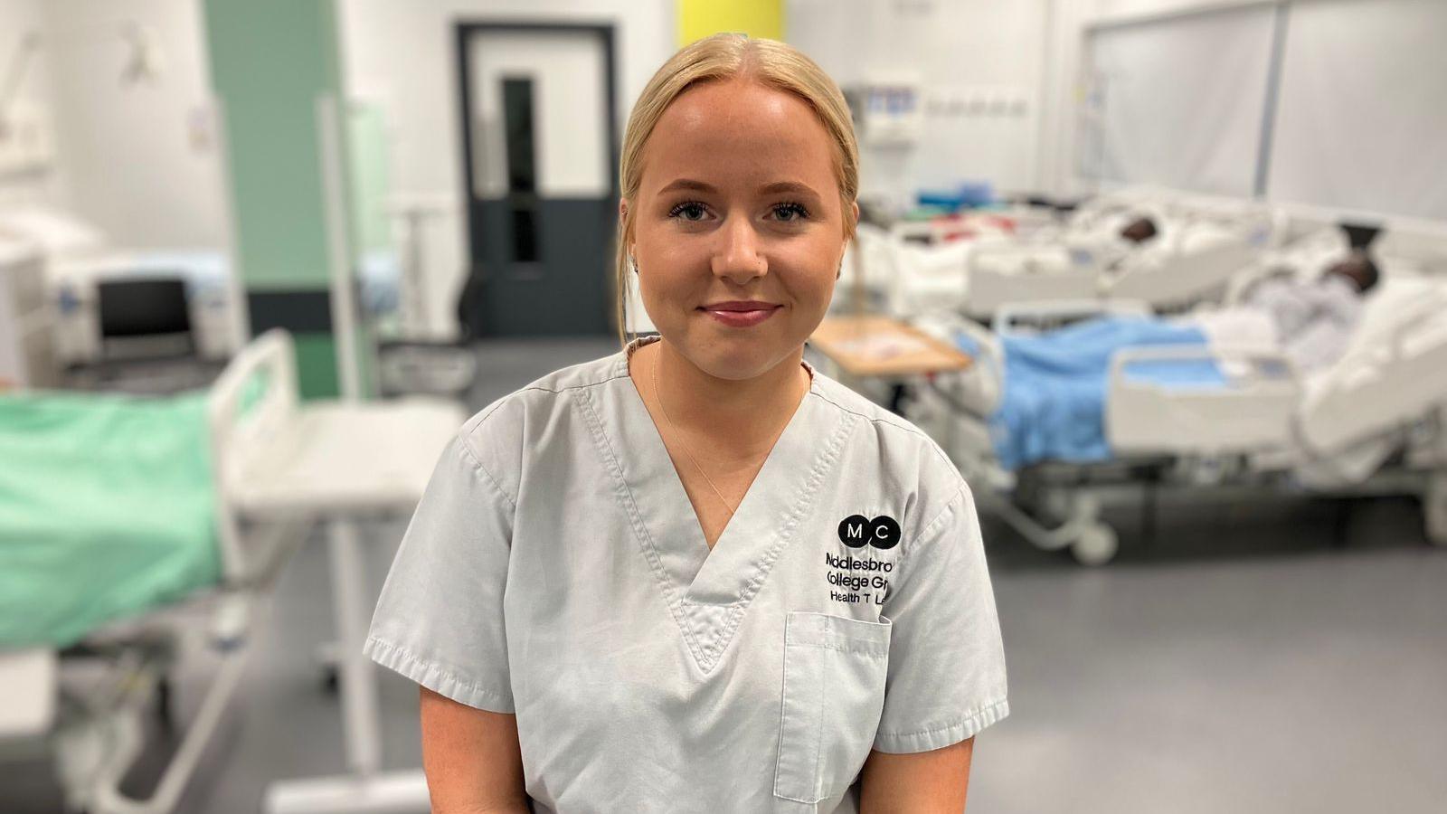 Freya has blonde hair tied back and wears a uniform with her college branding. Behind her, out of focus, is a mock-up hospital room in a college, with mannequins lying in hospital beds.